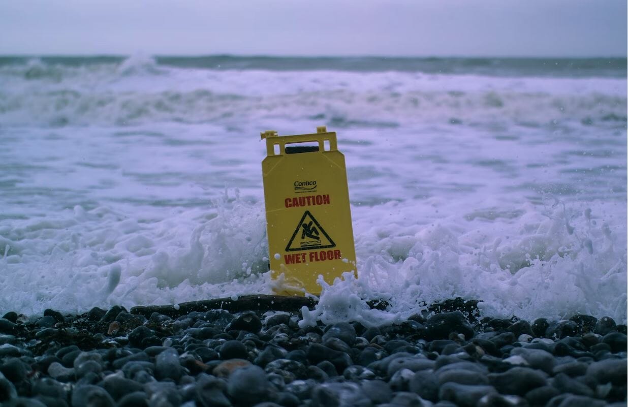 Un panneau attention sol mouillé est posé sur la plage et se fait recouvrir par les vagues. On peut l'associer aux difficultés que traverse YiedNodes dans ce bearmarket.